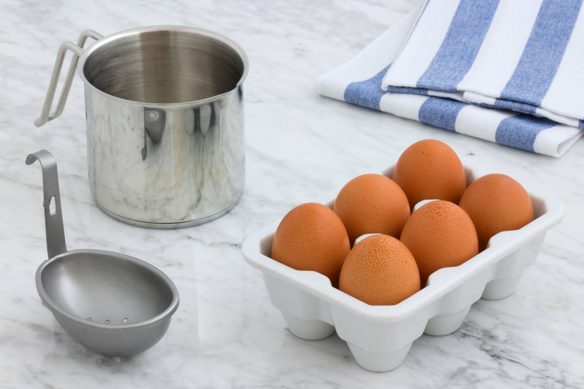 microwave egg poacher tool equipment needed showing stainless steel poacher cup with vented lid, silicone base tray, and measuring spoon arranged neatly on granite countertop